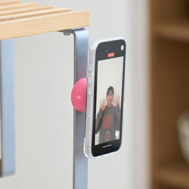 ピンクの磁石で金属製の棚にスマートフォンが固定され、画面には笑顔の女性が映っています。動画撮影やビデオ通話に便利なガジェットの利用シーンを示しています。