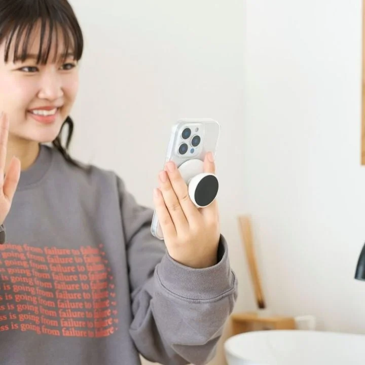 笑顔の若い女性が、スマホグリップを取り付けたスマートフォンを手に持っています。ビデオ通話をしているか、自撮りをしているような様子で、カジュアルな雰囲気です。