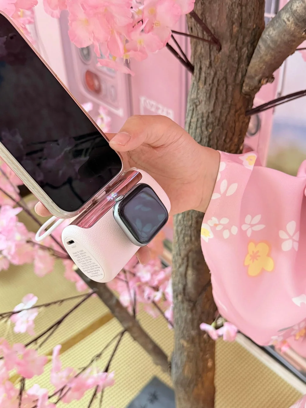 桜の装飾がある空間で、ピンク色の着物を着た人物がスマートフォンとスマートウォッチ用のポータブル充電器を持っている
