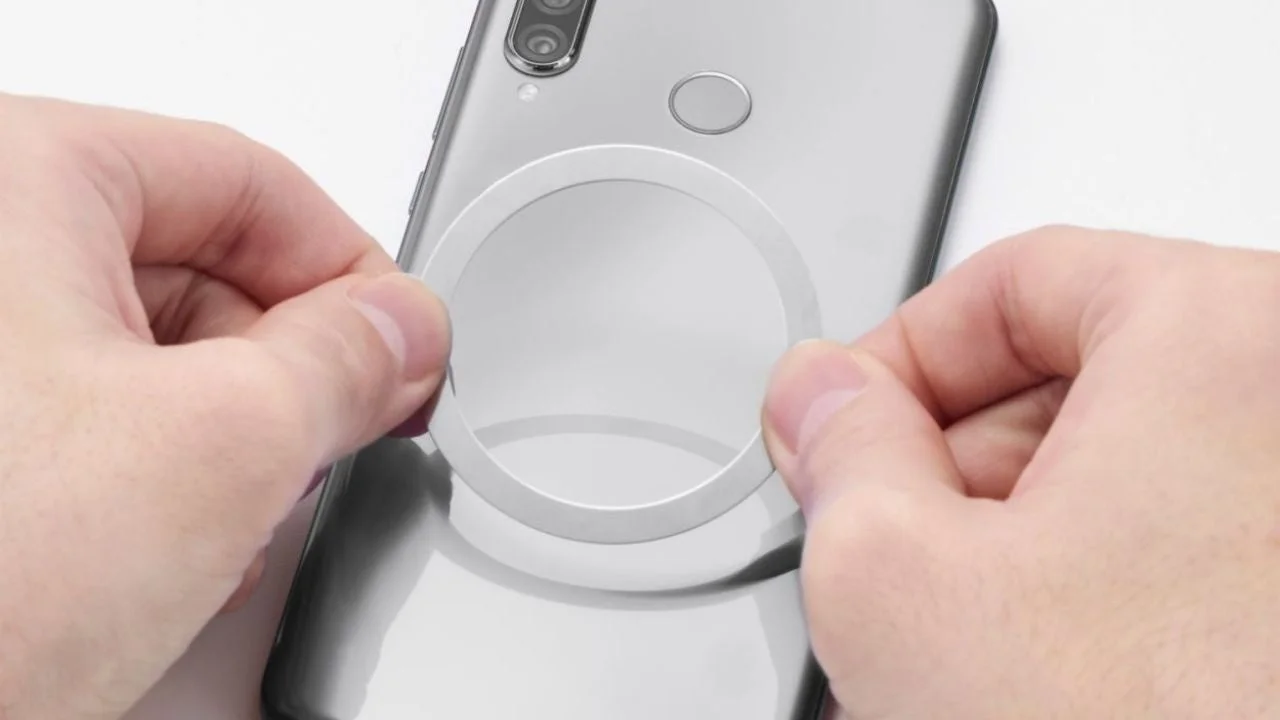 スマートフォンの背面に、円形の白いリング状のアクセサリーを貼り付けようとしている様子です。両手がリングをスマートフォンのカメラと指紋センサーの下あたりに位置合わせています。